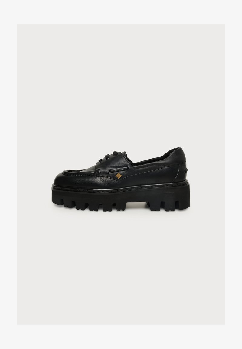 Mocassin en cuir noir avec une semelle épaisse et robuste et un petit emblème doré sur le côté, visible de l'extérieur sur un fond uni.