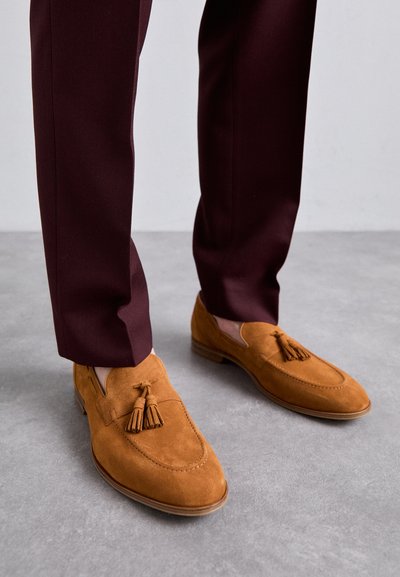 Tan suede loafers with tassel detail, rounded toe, and stitched accents, paired with burgundy tailored pants. Background is gray.
