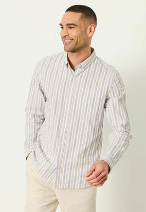 Lächelnder Mann, der ein beige-blau-braun vertikal gestreiftes langärmeliges Hemd mit beigen Hosen trägt und vor einem hellen Hintergrund steht.