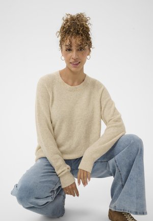 Beige pleteno pulover z rebrastimi manšetami, okroglo ovratnikom in mehkobo. V kombinaciji s svetlo modrimi širokimi hlačami in rjavimi čevlji na vezalke.