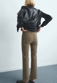 Giacca corta in pelle nera con orlo a coste, abbinata a pantaloni svasati di velluto a coste marrone chiaro e scarpe slip-on nere. Sfondo semplice.