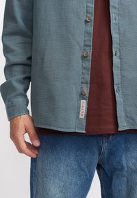 Lichtblauwe button-up shirt van een textuurachtige stof, met een bruine knopen en een "REVOLUTION" label. Daaronder is een bordeauxrood shirt, gecombineerd met denim jeans.