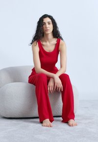 Γυναίκα με μακριά σγουρά μαλλιά που φοράει κόκκινο αμάνικο τοπ και παντελόνι, καθισμένη ξυπόλητη σε μια υφασμάτινη γκρι καρέκλα μπροστά σε απλό φόντο.