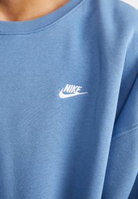 Kék Nike pulóver, kerek nyakkal, kis fehér logóval a mellkason. Simább textúra és laza kialakítás.