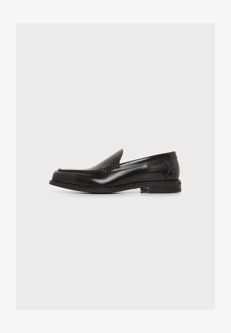 Mocassin en cuir noir à enfiler avec un petit talon et des détails cousus, étiquette latérale, sur fond blanc uni.