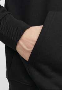 Schwarzer Pullover mit Rippbündchen und einer frontalen Tasche, mit strukturiertem Stoff, der an den Ärmeln und Taschennähten sichtbar ist.