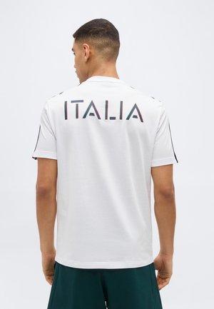 Mand iført en hvid kortærmet skjorte med "ITALIA" trykt med store bogstaver på ryggen, kombineret med mørkegrønne shorts.