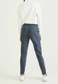 Person som bär en vit sweatshirt, marinblå tapered byxor och vita Reebok-sneakers, står mot en neutral ljus bakgrund, sett från baksidan.