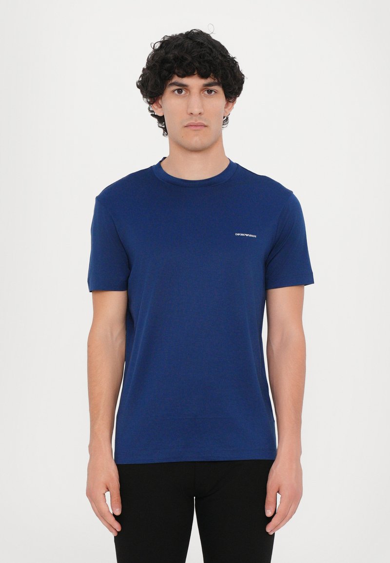 Tamsiai mėlynas apvalios apykaklės T-maišelis, pagamintas iš medvilnės, turi trumpas rankoves ir mažą prekės ženklo etiketę išsiuvinėtą kairėje krūtinės pusėje.