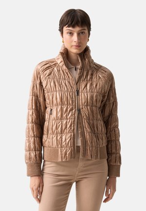 Frau trägt eine beige gesteppte Jacke mit hohem Kragen, Reißverschluss und gerippten Bündchen, kombiniert mit beigen Hosen vor einfachem Hintergrund.