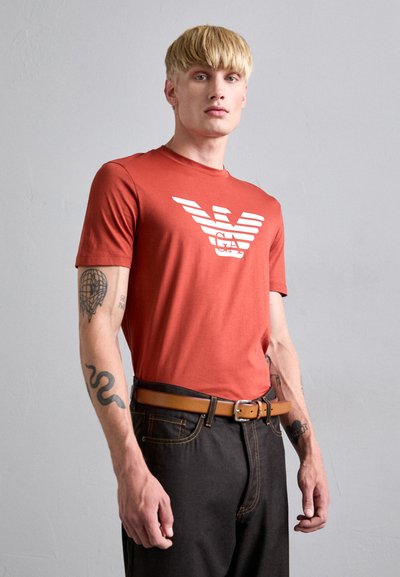 Men's red cotton t-shirt with white graphic print, featuring short sleeves and a crew neck, paired with dark jeans and a brown belt.