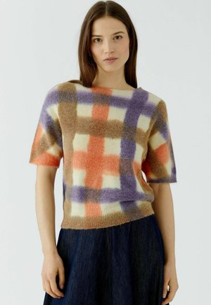 Jonge vrouw met steil bruin haar draagt een kortemouwige, zachte trui met een meerkleurig geruit patroon en een donkerblauwe rok tegen een witte achtergrond.