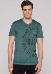Camiseta de rayas verde azulado con escote en V, que presenta texto y gráficos impresos en negro en el frente, mangas cortas y un corte relajado.