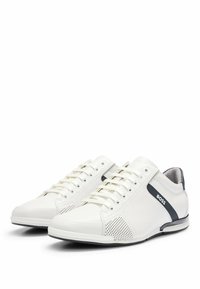 Zapatillas de cuero blanco con perforaciones, una franja de acento en color azul marino y suela de goma. Diseño con cordones y textura suave.