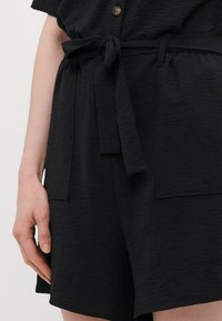 Svarte stoffshorts med strukturert overflate, belte med knyting i livet og to påsydde lommer foran. Knappebeslag øverst.