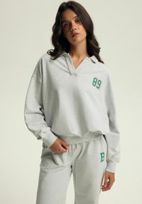Jasnoszara bluza z kołnierzykiem, z zielono-różowym nadrukiem "89" na przodzie. Materiał z mieszanki bawełny, luźny krój oraz ściągacze przy mankietach.
