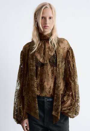 Durchsichtige braune Spitzenbluse mit hohem Kragen, gepufften Ärmeln und floralen Mustern, über einem schwarzen BH getragen, kombiniert mit dunklen Hosen und einem Gürtel.