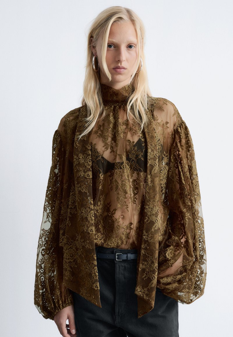 Durchsichtige braune Spitzenbluse mit hohem Kragen, gepufften Ärmeln und floralen Mustern, über einem schwarzen BH getragen, kombiniert mit dunklen Hosen und einem Gürtel.