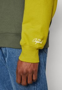 Felpa verde oliva e giallo brillante con polsino a coste. Presenta un testo ricamato bianco che recita "The Original" sulla manica gialla.