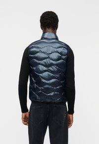 Άνδρας που φοράει γυαλιστερό, σκούρο μπλε, χωρίς μανίκια, καπιτονέ γιλέκο πάνω από μαύρο μακρυμάνικο πουκάμισο και μαύρα τζιν, γυρισμένος με την πλάτη προς το απλό φόντο.