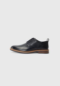 Zapato de cuero negro con punta redonda, diseño con cordones, un pequeño lazo en la parte trasera y un acento de suela marrón claro. Textura suave en toda la superficie.
