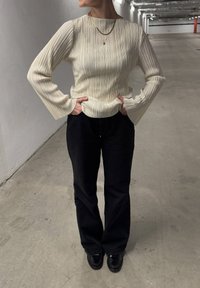 Lys beige plisseret langærmet top med bådudskæring, parret med sorte flagrende bukser og sorte sko. Teksturdetaljer er synlige.