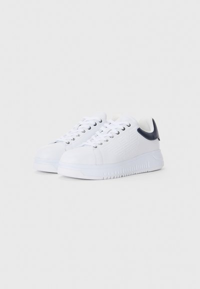 Hvite lave sneakers med preget logo på sidene og mørkeblå hælkonturer, vist i trekvartutsikt på en enkel bakgrunn.