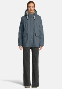 Vrouw in een blauwe gevoerde jas met knopen en voorzakken, gecombineerd met donkere wijde broek en lichte schoenen, staand tegen een witte achtergrond.