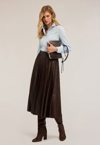 Camisa de botones azul claro, falda de imitación de cuero marrón con pliegues y botas de tacón alto de serpiente marrón. Sosteniendo un bolso oscuro con acentos azules.