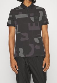 Polo à manches courtes noir avec motifs abstraits en blocs gris, patte de boutonnage et une poche poitrine ornée d'un logo carré noir.