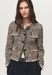 Giacca in tweed con un motivo testurizzato marrone e rosa, caratterizzata da bottoni neri, bordi sfrangiati e due tasche frontali, abbinata a una maglia bianca.