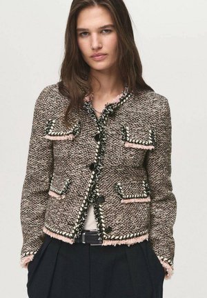 Giacca in tweed con un motivo testurizzato marrone e rosa, caratterizzata da bottoni neri, bordi sfrangiati e due tasche frontali, abbinata a una maglia bianca.