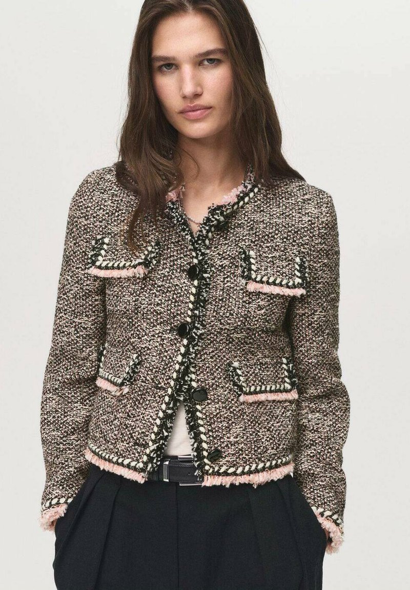 Giacca in tweed con un motivo testurizzato marrone e rosa, caratterizzata da bottoni neri, bordi sfrangiati e due tasche frontali, abbinata a una maglia bianca.