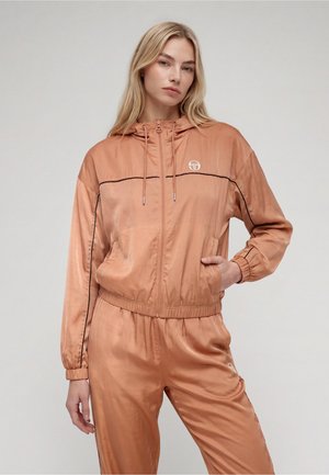 Femme portant un survêtement brillant couleur pêche avec passepoil noir et capuche, debout avec une main dans la poche devant un fond neutre.