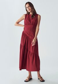 Rdeča brezrokavna maxi obleka z nabranimi pasovi, zaobljenim ovratnikom in prilegajočim pasom. Nosena z črnimi sandali z zlatim poudarkom.