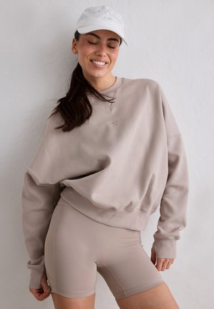 Kvinde med lukkede øjne smilende, iført beige oversized sweatshirt, matchende biker shorts og hvid baseballkasket mod en ensfarvet væg.