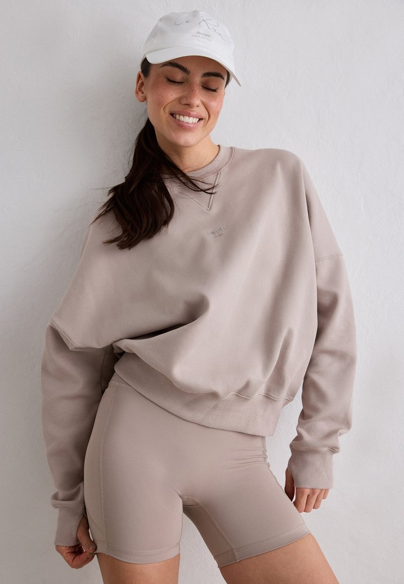 Femme aux yeux fermés souriant, portant un sweat-shirt beige surdimensionné, un short de cycliste assorti et une casquette de baseball blanche devant un mur uni.