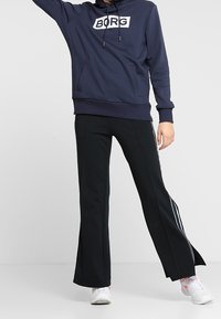 Sudadera azul marino con el logo "BORG" en un parche blanco, combinada con pantalones acampanados negros que tienen franjas laterales blancas, usados con zapatillas blancas.