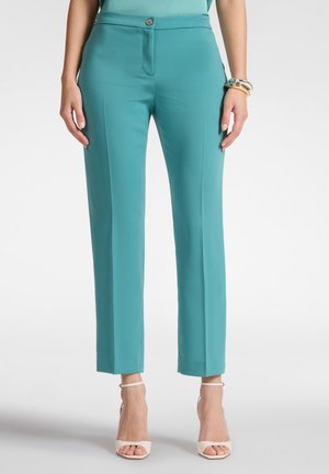 Vrouw draagt turquoise op maat gemaakte, enkellange broek met voorplooi, gecombineerd met beige open-toe schoenen en gouden armbanden om haar rechterpols.