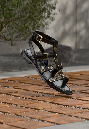 Czarna skórzana sandał gladiator z ozdobnymi złotymi ćwiekami i klamrami zawieszona nad drewnianą, kafelkowaną powierzchnią na zewnątrz.