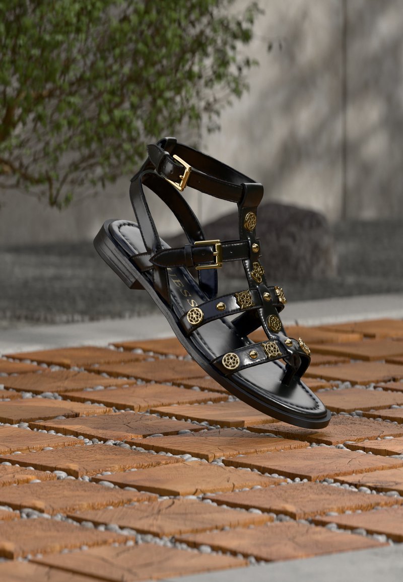 Μαύρο δερμάτινο σανδάλι γλαδιaτώρα με χρυσά διακοσμητικά καρφιά και αγκράφες, κρεμασμένο πάνω από ξύλινη πλακόστρωτη επιφάνεια σε εξωτερικό χώρο.