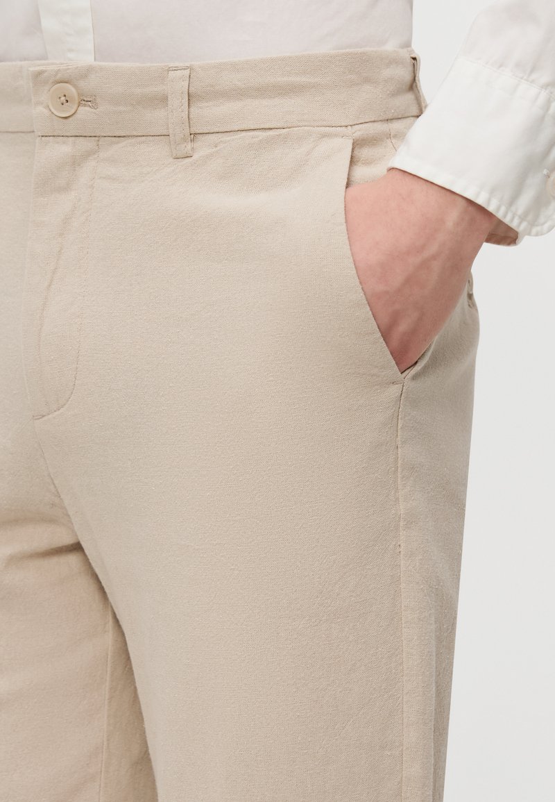 Pantalon beige clair en tissu texturé, avec une fermeture à bouton, une poche latérale et une coupe ajustée. Coutures visibles le long des coutures.