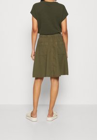 Falda verde oliva con diseño acampanado, cintura elástica y dos bolsillos traseros. Combinada con un top oscuro y zapatos claros.