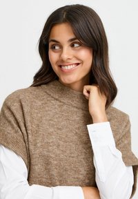 Beige strikket turtleneck sweater med korte ærmer, der har en blød tekstur og en afslappet pasform, lagt over en hvid skjorte med manchetter.