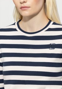 Striped sweater featuring navy blue and cream colors, round neckline, ribbed texture, and a small embroidered logo on the chest.