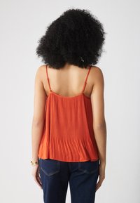 Débardeur plissé rouge avec de fines bretelles, présentant une texture lisse. Assorti à un jean bleu foncé, affichant une coupe décontractée.