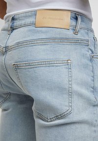 Gai zils denims ar aizmugures kabatu un brūnu ādas etiķeti "2V PREMIUM", valkāti ar baltu ribotu blūzi.