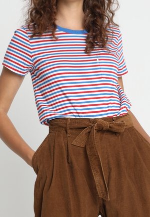 Vrouw draagt een rood, wit en blauw gestreept t-shirt, ingestopt in een bruin hooggetailleerd corduroybroek met een gestrikte riem.