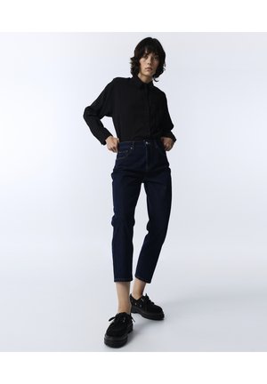 Person trägt schwarzes Hemd mit Knöpfen, dunkelblaue kurze Jeans und schwarze Plateauschuhe und steht vor weißem Hintergrund.