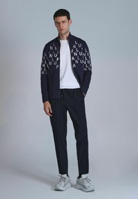 Marine ritsjack met witte tekstaccenten op de mouwen, gecombineerd met een wit T-shirt en donkere broek. Model draagt grijze sneakers.